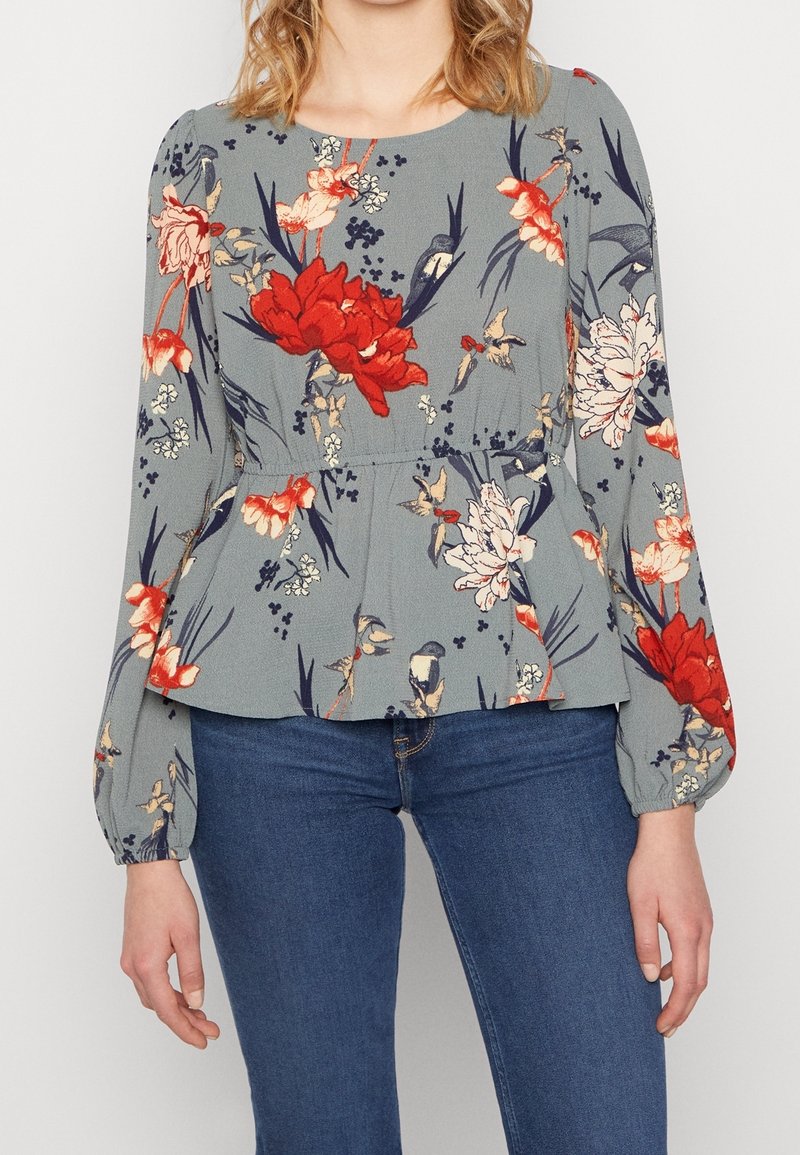 Graue Bluse mit Blumenmuster in Rot und Weiß, mit langen Ärmeln und gerafftem Taillenbereich, aus leichtem Stoff gefertigt.