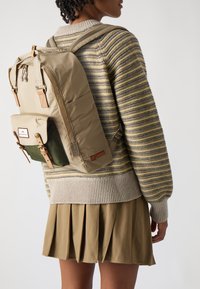 Beige backpack with green accents, featuring padded straps, a front pocket, and leather detailing. Worn over a striped sweater and pleated skirt.