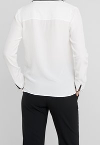 Witte langwerpige blouse met zwarte bies langs de kraag en manchetten, gemaakt van een soepele, lichte stof en met een relaxte pasvorm.