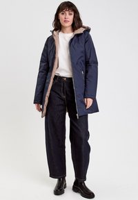 Manteau rembourré bleu marine avec une doublure beige, doté d'une capuche, d'une fermeture éclair à l'avant et de poches latérales. Associé à un jean large noir et à des bottines noires.