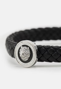 Svart fletteleddbånd i lær med en sølvfarget løvehode-klaff og en sirkulær metallaccent gravert med "GUESS" og "LOS ANGELES."