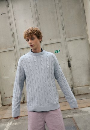 Light blue cable-knit sweater with long sleeves and a ribbed neckline, worn over a black top, paired with light pink shorts.