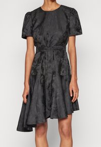 Robe noire avec une finition texturée, manches bouffantes courtes, taille cintrée et jupe évasée. Présente une ceinture nouée en accent et une longueur mi-genou.