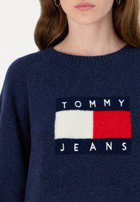 Maglione lavorato a maglia blu navy con un logo rettangolare con "TOMMY" in bianco, un rettangolo bianco e un rettangolo rosso sopra "JEANS". Tessuto morbido.