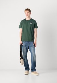 Grøn bomulds T-shirt med korte ærmer og et hvidt logo, parret med lyseblå jeans og beige sko samt en lille crossbody taske.