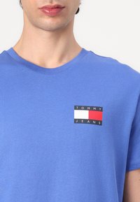 Camiseta de algodón azul con un pequeño parche de logo que dice "TOMMY" en blanco, "JEANS" en negro, y rectángulos rojos y blancos, con cuello redondo.