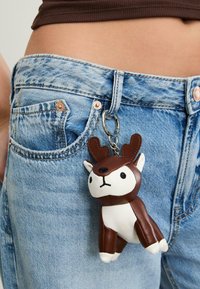 Portachiavi in peluche marrone e bianco a forma di un cervo stilizzato con corna, attaccato a un anello metallico argento, appeso a dei jeans in denim azzurro chiaro.