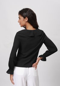 Schwarzes Langarmoberteil mit großem Kragen, strukturiertem Stoff und voluminösen Manschetten. Getragen mit weißen Hosen, die über Gesäßtaschen verfügen.