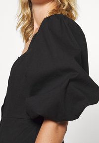 Czarna sukienka z bufiastymi rękawami wykonana z fakturowanej tkaniny, z kwadratowym dekoltem i dopasowanym korpusem, podkreślająca miękki, obszerny krój rękawów.