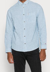 Vaaleansininen ja valkoinen raidallinen pitkähihainen paita, jossa on napituskaulus, rintatasku ja metallinappulat; sisältää brändättyä hihansuutekstiä.