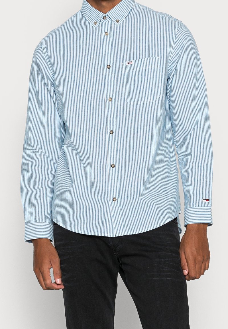 Vaaleansininen ja valkoinen raidallinen pitkähihainen paita, jossa on napituskaulus, rintatasku ja metallinappulat; sisältää brändättyä hihansuutekstiä.