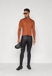 Camisola de ciclismo justa, de manga comprida, na cor ferrugem com acentos cinzentos, combinada com calças justas pretas almofadadas. Detalhes adicionais incluem um capacete.