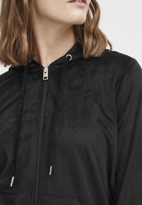 Hoodie noir zippé en tissu doux, doté d'une capuche, de cordons de serrage et d'accents de fermeture éclair métalliques. Texture lisse avec une légère brillance.
