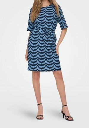 Femme portant une robe aux motifs bleu et noir, longueur genou, manches trois-quarts, et des sandales noires à talons hauts, debout avec une main sur la hanche.
