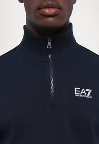 Marineblå zip-neck genser i glatt stoff. Har glitrende sølvglidelås og brodert "EA7 Emporio Armani"-logo på brystet.