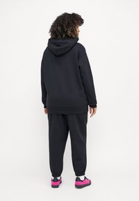 Černá mikina s kapucí a odpovídající tepláky vyrobené z měkké tkaniny. Mikina má šňůrku; tepláky mají elastické manžety. Odvážné růžové boty jako akcent.