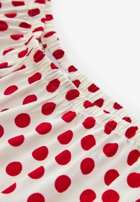 Biela látka s červenými bodkami, s nariasenou elastickou časťou okolo pásu. Textúra je hladká s ľahkým vzhľadom.