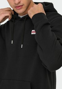 Zwarte hoodie van een katoenmix, met een buidelzak, verstelbare koordjes en een klein rood-wit logo-op een patch op de borst.