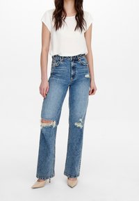 Jean bleu taille haute avec déchirures usées, coupe ample et ourlets bruts, associé à un top blanc à manches courtes et des chaussures pointues beiges.