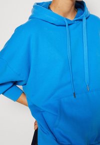 Felpa oversize blu in cotone, con tasca frontale, cordini con punte metalliche e polsini a coste. Texture morbida.