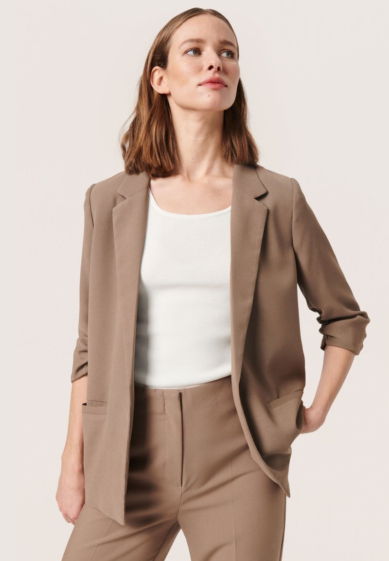 Hnědý blazer se strukturovaným designem, vybavený klopami a ohrnutými rukávy. Nosený přes bílé tričko, doplněný o odpovídající kalhoty.