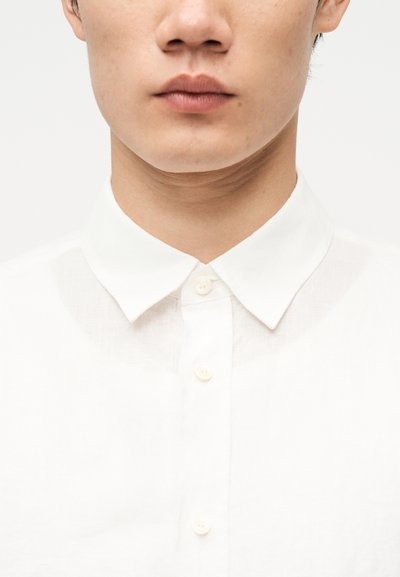 Κοντινό πλάνο του κάτω μέρους του προσώπου και του λαιμού ενός ατόμου που φοράει ένα λευκό πουκάμισο με κουμπιά και γιακά, μπροστά από ένα απλό φόντο.