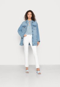 Denim jakna svijetloplave boje, širokog kroja. Plava bluzica sa teksturom i detaljem u obliku ključa. Bijele traperice ravnog kroja i svijetloplave sandale s visokom potpeticom.