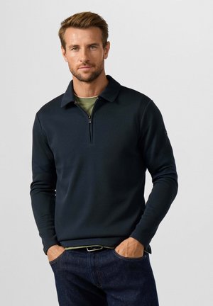 Man draagt een donkerblauwe trui met een kwart rits over een groen shirt en blauwe jeans, staand met handen in de zakken tegen een effen achtergrond.