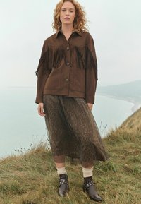 Brown suede jacket with black fringe details, paired with a sheer, leopard-print skirt, cream socks, and black ankle boots with silver accents.