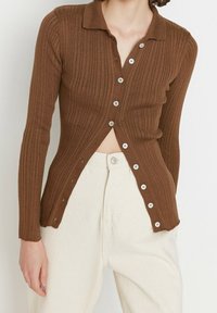 Brun ribbad cardigan med krage, långärmade ärmar och vita knappar framtill, i kombination med ljusa byxor.