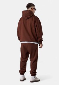 Sweat à capuche marron avec un pantalon de jogging assorti, affichant une coupe décontractée, des poignets côtelés et une texture douce. Porté avec des baskets de couleur claire.