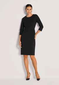 Zwarte knielange jurk met driekwartmouwen, met een getailleerd silhouet en een subtiel gestructureerd ontwerp. Gecombineerd met hoge hakken.