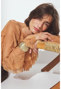 Woman with short brown hair resting head and hands on a wooden chair wrapped in woven rattan, wearing a peach lace blouse and gold bracelet.
