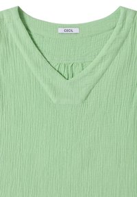 Haut en tissu texturé vert clair avec un col en V et un détail froncé sous le col portant l'étiquette « Cecil ».