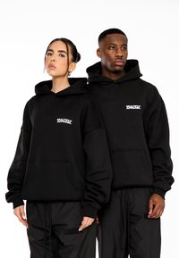 Sudaderas negras con bolsillos frontales, que presentan un logo blanco de "MULTIPLY APPAREL". Modeladas por dos personas, combinadas con pantalones cargo negros.