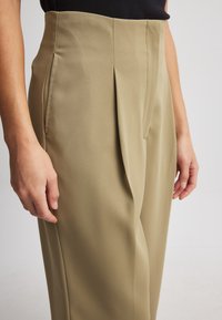 Khaški nabrani hlače z visokim pasom, izdelane iz gladkega materiala, z bočnimi žepi in oblikovanim krojem.
