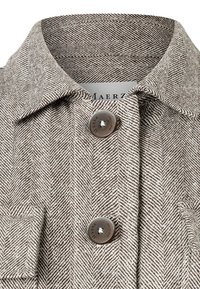 Cuello de camisa con patrón de espiga en tonos beige y marrón, con dos botones marrones y un bolsillo en el pecho. Etiqueta de la marca visible en el interior del cuello.