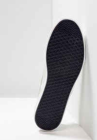 Sneakersula i strukturerad svart gummi med ett geometriskt mönster, kantad med en slät vit mellansula. Ljust beige ovandel synlig vid kanten.