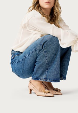 Donna accovacciata che indossa scarpe slingback beige chiaro con tacco e fibbie decorative, jeans blu e una camicetta bianca trasparente.