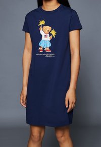 Navy bomullst-shirtklänning med en grafik av en björn i vit skjorta och blå kjol, som håller gula pom-poms. Avslappnad passform, korta ärmar.