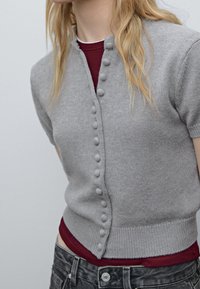 Cardigan grigio lavorato a maglia con scollo rotondo e maniche corte, caratterizzato da una fila di bottoni testurizzati sul davanti e vita aderente.