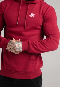 Felpa con cappuccio rossa con tasca frontale, polsini e orlo a costine. Logo bianco ricamato sul petto. Realizzata in tessuto morbido ed elastico.