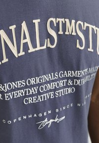 Navyblauwe katoenen t-shirt met grote witte tekst en een handtekening. De tekst bevat de merknaam en details over comfort en duurzaamheid.