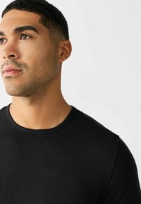 T-shirt nera con scollo rotondo in tessuto liscio, dal design aderente e maniche corte. Primo piano sullo scollo e dettagli della parte superiore del corpo.