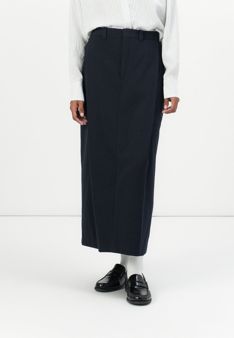 Jupe longue navy au cut straight, avec des plis sur le devant et des passants de ceinture. Associée à des mocassins noirs et des chaussettes blanches.