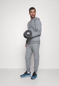 Grauer sportlicher Kapuzenpullover und Jogginghose, hält eine schwarze Gewichtsscheibe. Die Sneakers sind blau mit schwarzen Akzenten. Das Outfit hat eine lockere Passform.