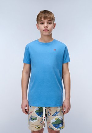 Ragazzo che indossa una t-shirt a maniche corte azzurro chiaro e pantaloncini beige con stampe colorate a tema viaggio e oceano, in piedi davanti a uno sfondo semplice.