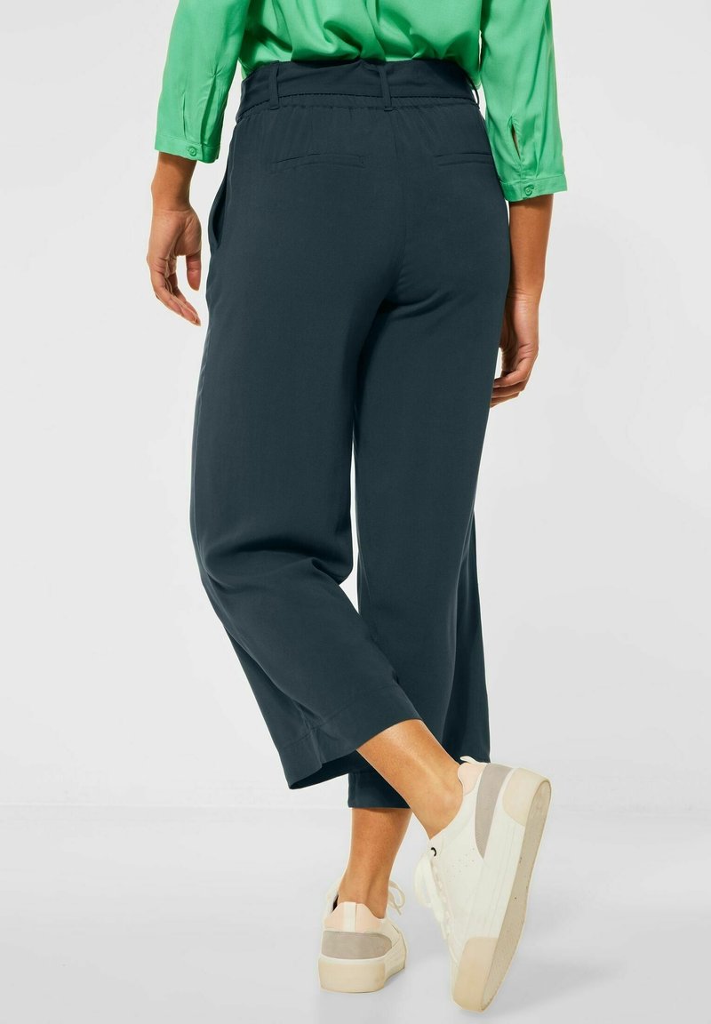 Personne portant un pantalon court foncé, une chemise verte à manches longues et des baskets blanches, debout avec une jambe croisée derrière l'autre.
