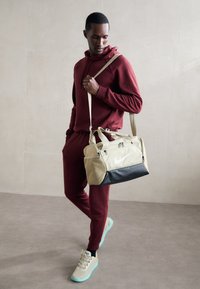 Sac de sport Nike beige avec une base noire, doté de compartiments zippés et d'un grand logo. L'homme porte un survêtement bordeaux.
