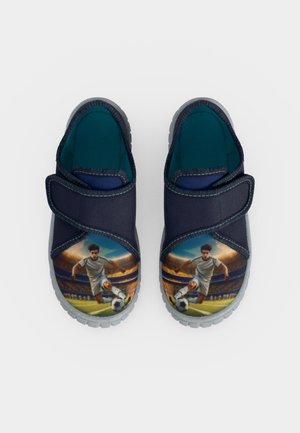 Par de zapatos infantiles azul marino con tiras de velcro, cada uno mostrando a un futbolista chutando un balón en un estadio sobre la puntera.